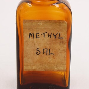 May include: Amber-colored glass bottle with a rectangular label. The label reads "METHYL SAL" in handwritten black ink. The bottle has a square shape with rounded edges and a small neck.