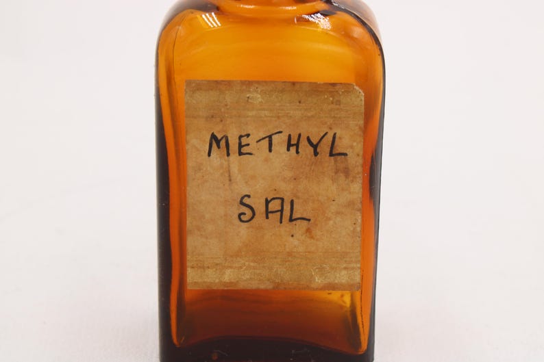 May include: Amber-colored glass bottle with a rectangular label. The label reads "METHYL SAL" in handwritten black ink. The bottle has a square shape with rounded edges and a small neck.