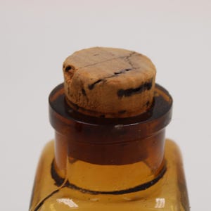 May include: Close-up of a vintage amber glass bottle with a cork stopper. The bottle has a square shape and a dark brown neck. The cork is light brown with dark cracks.