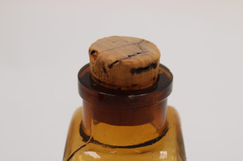 May include: Close-up of a vintage amber glass bottle with a cork stopper. The bottle has a square shape and a dark brown neck. The cork is light brown with dark cracks.