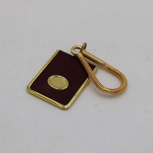 May include: A rectangular key fob with a burgundy leather surface and a gold-colored frame. It features a gold oval emblem and a coiled gold key ring. The key fob is a classic accessory.