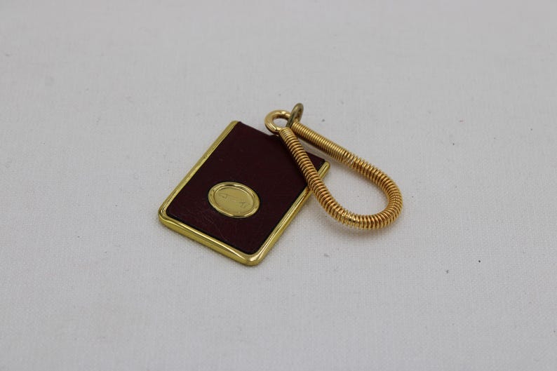 May include: A rectangular key fob with a burgundy leather surface and a gold-colored frame. It features a gold oval emblem and a coiled gold key ring. The key fob is a classic accessory.
