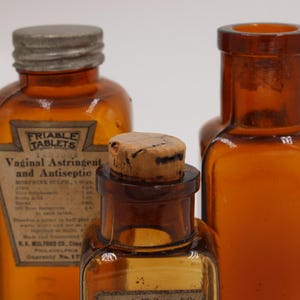 May include: Three antique amber glass bottles. The bottles have different shapes and sizes, with one labeled "Friable Tablets Vaginal Astringent and Antiseptic." The bottles have metal and cork stoppers. The background is plain white.