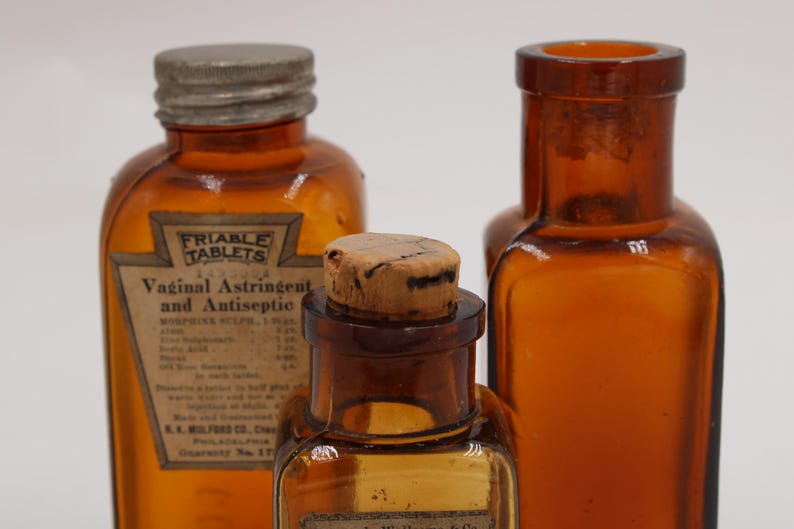 May include: Three antique amber glass bottles. The bottles have different shapes and sizes, with one labeled "Friable Tablets Vaginal Astringent and Antiseptic." The bottles have metal and cork stoppers. The background is plain white.
