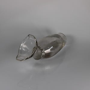 May include: A clear glass pitcher with a spout, lying on its side. The pitcher has a rounded body and a flared spout. The glass is transparent, allowing a view of the interior. The pitcher is resting on a light gray surface.