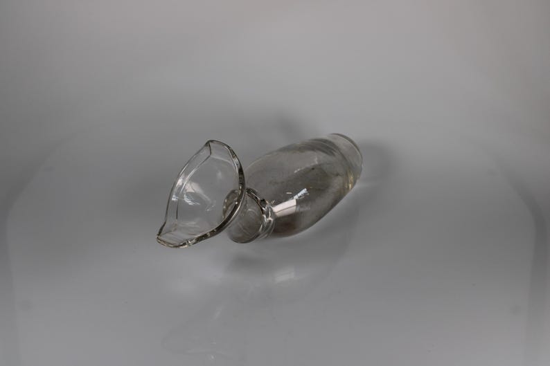 May include: A clear glass pitcher with a spout, lying on its side. The pitcher has a rounded body and a flared spout. The glass is transparent, allowing a view of the interior. The pitcher is resting on a light gray surface.