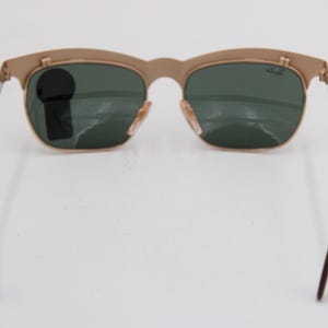 May include: Gold-tone sunglasses with green lenses. The sunglasses have a classic design with a curved top frame and thin metal arms. The lenses are a dark green color, and the frame is a light gold color. The arms have dark brown tips.