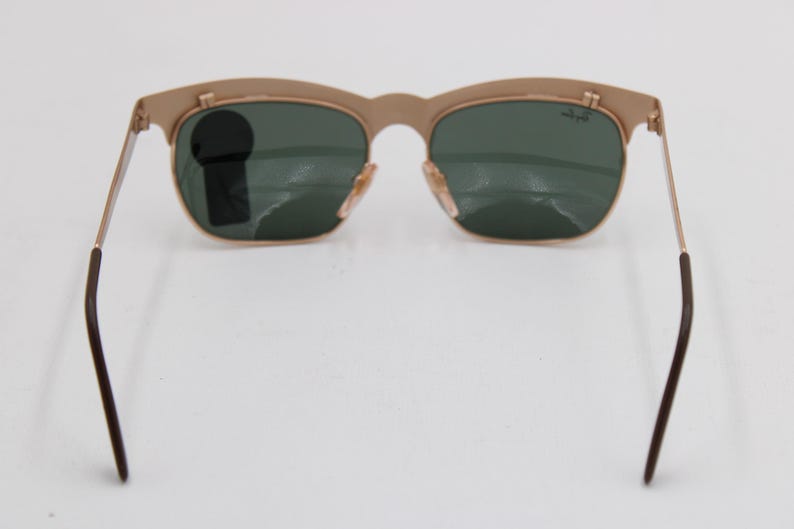 May include: Gold-tone sunglasses with green lenses. The sunglasses have a classic design with a curved top frame and thin metal arms. The lenses are a dark green color, and the frame is a light gold color. The arms have dark brown tips.
