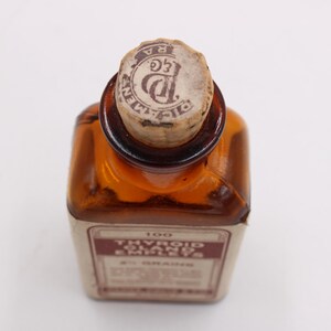 May include: An antique amber glass bottle with a cork stopper. The bottle has a rectangular shape and a label with text. The cork is stamped with a circular design. The label reads "Thyroid Glands" and "100 2 1/2 GRAINS".