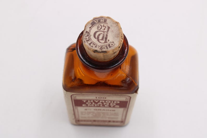 May include: An antique amber glass bottle with a cork stopper. The bottle has a rectangular shape and a label with text. The cork is stamped with a circular design. The label reads "Thyroid Glands" and "100 2 1/2 GRAINS".
