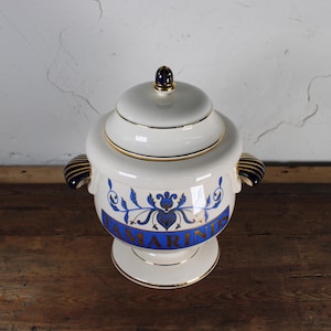 May include: A white ceramic jar with a lid, gold trim, and blue floral designs. The jar has two handles and the word "TAMARINDS" in blue lettering. The jar is on a pedestal base and sits on a wooden surface.