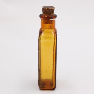 May include: An amber glass bottle with a cork stopper. The rectangular bottle has rounded corners and a textured surface. The cork is light brown. The bottle is empty and stands against a white background. The bottle is approximately 10 cm tall.