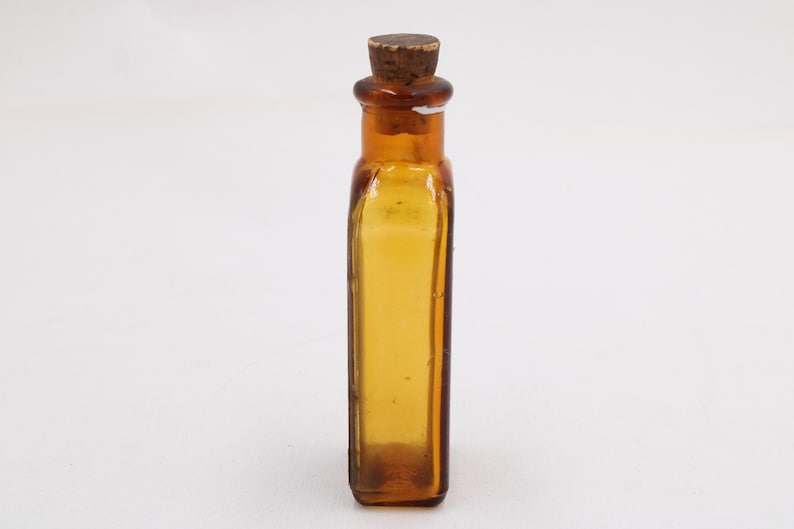 May include: An amber glass bottle with a cork stopper. The rectangular bottle has rounded corners and a textured surface. The cork is light brown. The bottle is empty and stands against a white background. The bottle is approximately 10 cm tall.