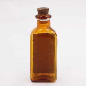 May include: An amber glass bottle with a cork stopper. The rectangular bottle has a label on the front, possibly an antique apothecary or medicine bottle. The bottle is empty and stands on a white background.