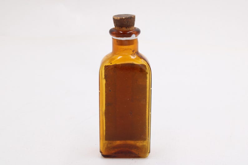 May include: An amber glass bottle with a cork stopper. The rectangular bottle has a label on the front, possibly an antique apothecary or medicine bottle. The bottle is empty and stands on a white background.