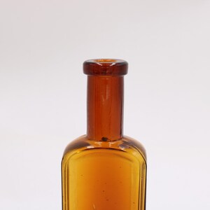 May include: Amber-colored glass bottle with a rectangular body and a long, cylindrical neck. The bottle has a rounded shoulder and a dark brown cap. The glass has a slight texture. The bottle is empty.