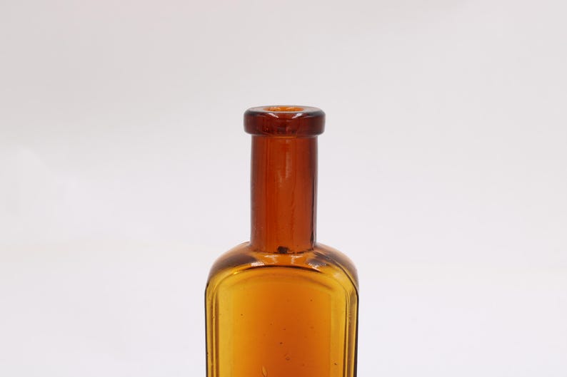May include: Amber-colored glass bottle with a rectangular body and a long, cylindrical neck. The bottle has a rounded shoulder and a dark brown cap. The glass has a slight texture. The bottle is empty.