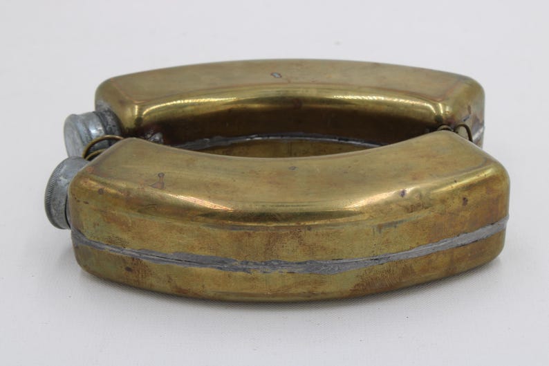 May include: A vintage brass flask with a curved, rectangular shape. The flask is made of brass with a tarnished, aged appearance. It has two screw-on caps and a silver-colored seam. The flask is likely used for carrying liquids.