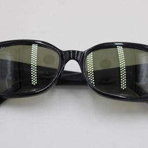 Puede incluir: Gafas de sol negras con montura cuadrada y lentes verdes. Las gafas de sol tienen un dise&ntilde;o cl&aacute;sico con un logotipo sutil en la esquina superior izquierda de la montura. Las lentes reflejan la luz.