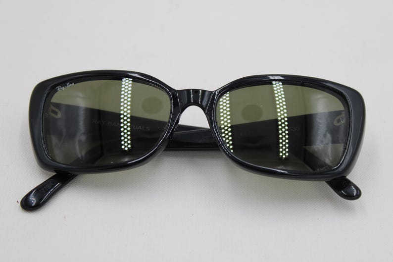 Puede incluir: Gafas de sol negras con montura cuadrada y lentes verdes. Las gafas de sol tienen un dise&ntilde;o cl&aacute;sico con un logotipo sutil en la esquina superior izquierda de la montura. Las lentes reflejan la luz.