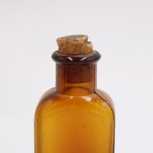 May include: Amber glass bottle with a cork stopper. The bottle has a rounded shape and a dark brown neck. The cork is a light brown color and appears to be made of wood. The bottle is empty and sits against a white background.