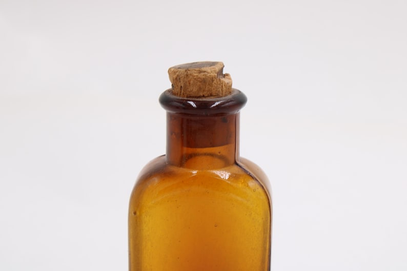 May include: Amber glass bottle with a cork stopper. The bottle has a rounded shape and a dark brown neck. The cork is a light brown color and appears to be made of wood. The bottle is empty and sits against a white background.