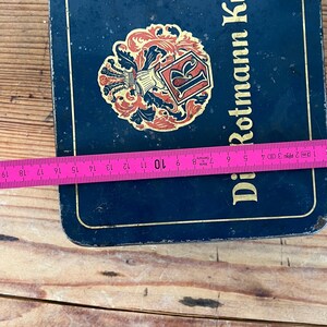 May include: A rectangular, dark blue metal box with the words "Di Rotmann Krone" in gold lettering. The box features a decorative emblem with red and gold accents. A measuring tape shows the box is approximately 7 inches wide.