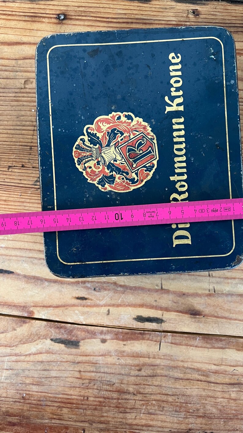 May include: A rectangular, dark blue metal box with the words "Di Rotmann Krone" in gold lettering. The box features a decorative emblem with red and gold accents. A measuring tape shows the box is approximately 7 inches wide.