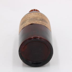 Puede incluir: Una botella de vidrio marr&oacute;n vintage con una etiqueta. La botella tiene una forma redondeada y un color &aacute;mbar oscuro. La etiqueta parece tener texto y es de color marr&oacute;n claro. La botella est&aacute; acostada de lado.