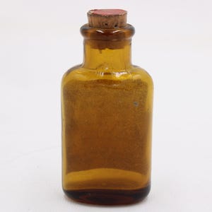 May include: Amber glass bottle with a cork stopper. The bottle has a rectangular body and a rounded neck. The cork is light brown with a red top. The bottle is empty and sits on a white background.