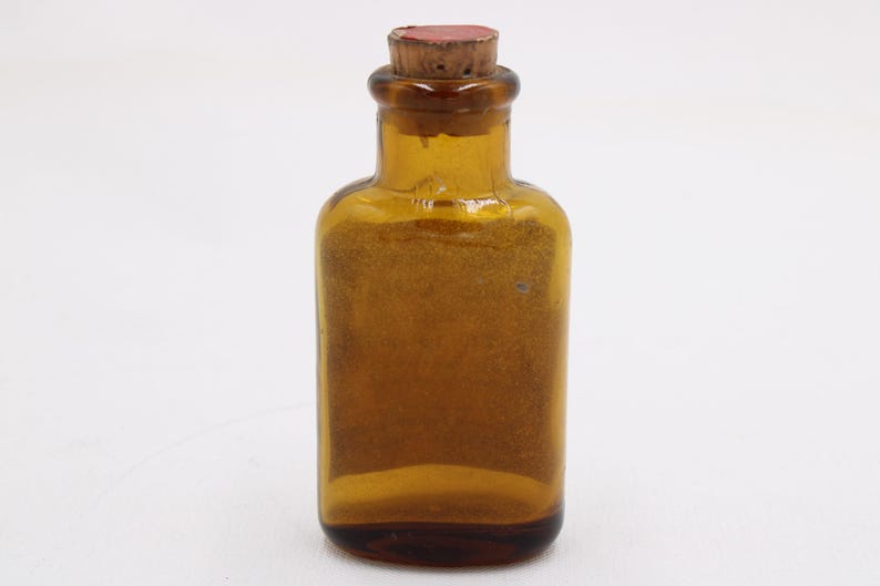 May include: Amber glass bottle with a cork stopper. The bottle has a rectangular body and a rounded neck. The cork is light brown with a red top. The bottle is empty and sits on a white background.