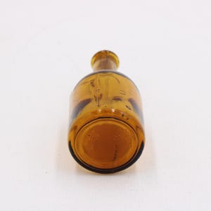 May include: Amber-colored glass bottle with a rounded body and a narrow neck. The bottle is lying on its side, showcasing its shape and color. The glass appears to be slightly textured.
