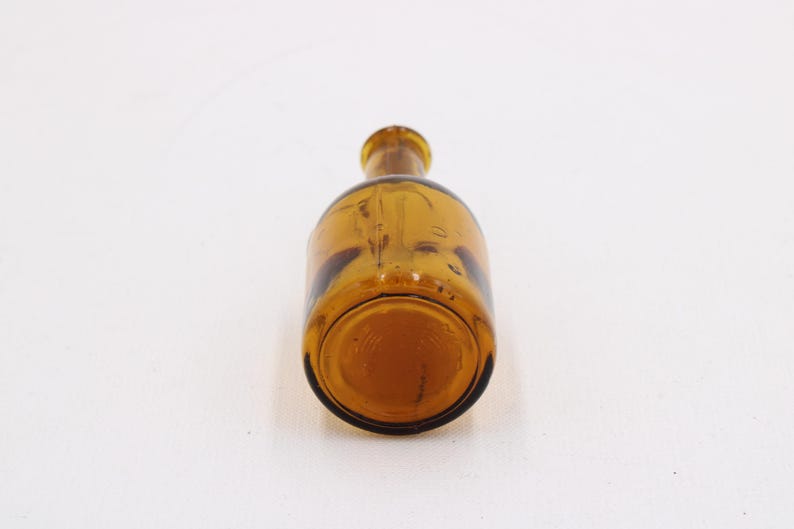 May include: Amber-colored glass bottle with a rounded body and a narrow neck. The bottle is lying on its side, showcasing its shape and color. The glass appears to be slightly textured.