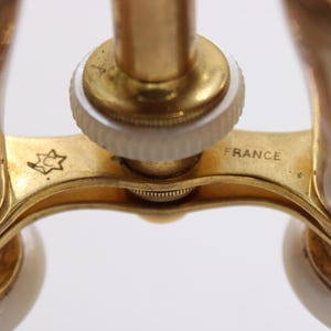 May include: Close-up of antique opera glasses with a gold-tone frame and iridescent mother-of-pearl accents. The word "FRANCE" is visible on the frame. A star-shaped emblem is also present.
