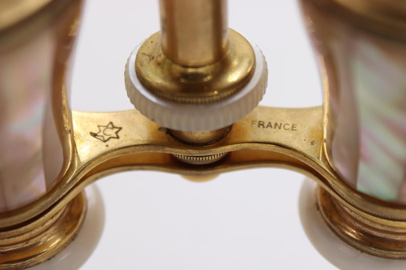 May include: Close-up of antique opera glasses with a gold-tone frame and iridescent mother-of-pearl accents. The word "FRANCE" is visible on the frame. A star-shaped emblem is also present.