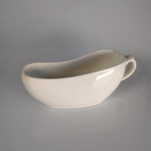Könnte beinhalten: Eine weiße, nierenförmige Sauciere mit Griff. Das Keramikboot hat eine glänzende Oberfläche und ist vor einem neutralen Hintergrund platziert. Die Sauciere ist zum Servieren von Saucen und Soßen bestimmt.