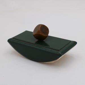 Puede incluir: Un secante de tinta verde oscuro con una base redondeada y un pequeño mango marrón en forma de cubo. El secante es rectangular y tiene una parte superior ligeramente curvada. El mango es de color marrón oscuro.