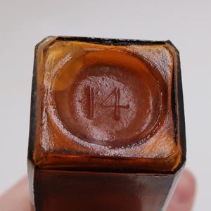 May include: Close-up of the bottom of a brown glass bottle. The number "14" is embossed in a circular indentation. The bottle has a square shape with a slightly rounded base.