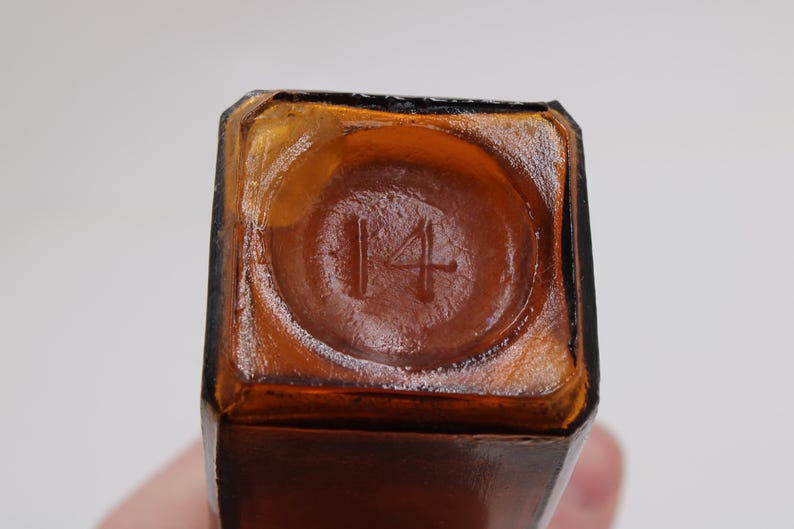 May include: Close-up of the bottom of a brown glass bottle. The number "14" is embossed in a circular indentation. The bottle has a square shape with a slightly rounded base.