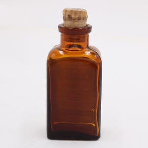 May include: A small, amber-colored glass bottle with a square base and a cork stopper. The bottle has a rectangular shape with rounded corners and a narrow neck. The cork is light brown and fits snugly in the bottle's opening. The bottle is empty.