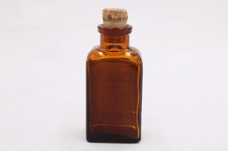 May include: A small, amber-colored glass bottle with a square base and a cork stopper. The bottle has a rectangular shape with rounded corners and a narrow neck. The cork is light brown and fits snugly in the bottle's opening. The bottle is empty.