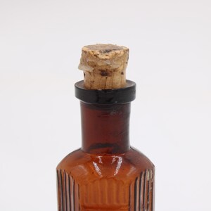 Puede incluir: Una peque&ntilde;a botella de vidrio de color &aacute;mbar con un tap&oacute;n de corcho. La botella tiene estr&iacute;as verticales y un anillo negro alrededor del cuello. El corcho es de color marr&oacute;n claro y parece estar hecho de material natural.