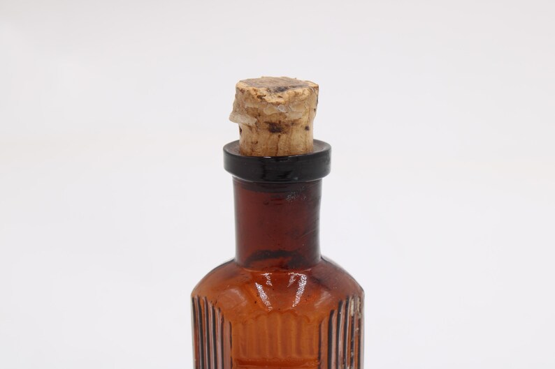 Puede incluir: Una peque&ntilde;a botella de vidrio de color &aacute;mbar con un tap&oacute;n de corcho. La botella tiene estr&iacute;as verticales y un anillo negro alrededor del cuello. El corcho es de color marr&oacute;n claro y parece estar hecho de material natural.