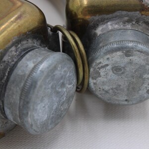 May include: Close-up of two antique brass containers with silver-colored screw-on lids. The containers are connected by a brass ring. The brass has a worn, aged appearance, with signs of oxidation and wear.