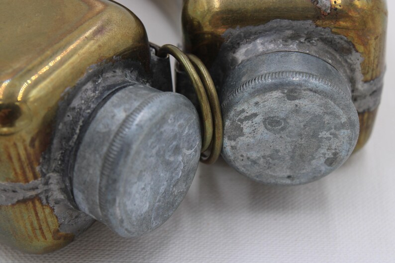 May include: Close-up of two antique brass containers with silver-colored screw-on lids. The containers are connected by a brass ring. The brass has a worn, aged appearance, with signs of oxidation and wear.