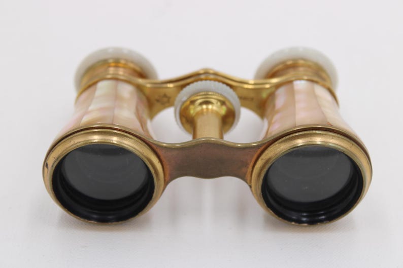 May include: Antique opera glasses with a gold-tone metal frame and mother-of-pearl accents. The lenses are black, and the binoculars have a central focus wheel. The design is ornate and vintage.