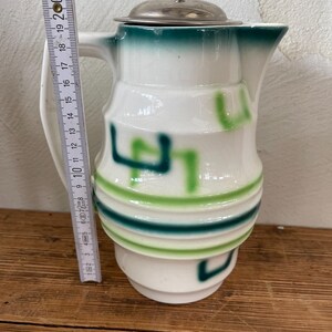 K&ouml;nnte beinhalten: Wei&szlig;er Keramikkrug mit silbernem Deckel und Griff. Der Krug hat gr&uuml;ne und hellgr&uuml;ne Akzente. Der Krug ist etwa 18 cm hoch.