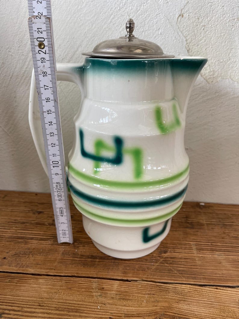 K&ouml;nnte beinhalten: Wei&szlig;er Keramikkrug mit silbernem Deckel und Griff. Der Krug hat gr&uuml;ne und hellgr&uuml;ne Akzente. Der Krug ist etwa 18 cm hoch.