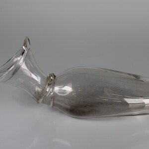 May include: A clear glass decanter with a flared, curved spout and a bulbous body. The decanter is lying on its side, showcasing its elegant shape. The glass appears to be slightly tinted, adding a subtle visual depth.
