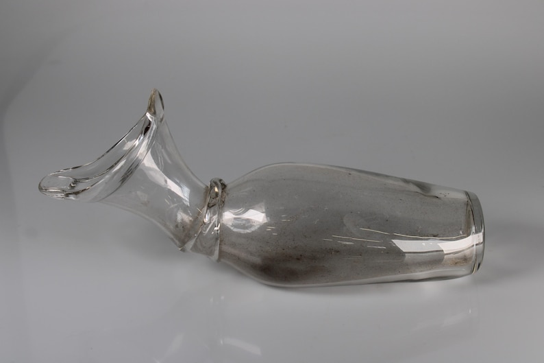 May include: A clear glass decanter with a flared, curved spout and a bulbous body. The decanter is lying on its side, showcasing its elegant shape. The glass appears to be slightly tinted, adding a subtle visual depth.